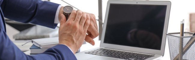 Profissional conferindo o relógio de pulso ao lado de um notebook, representando o controle de jornada e a gestão do banco de horas no desligamento.