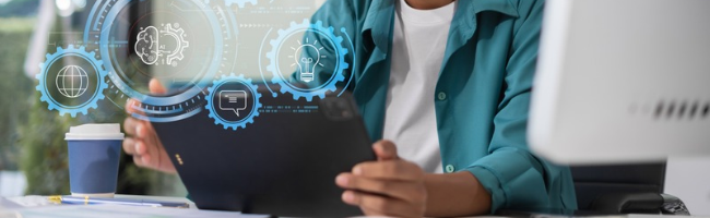 Pessoa de camisa verde-azulada usando um tablet em uma mesa de trabalho, cercada por ícones digitais holográficos em engrenagens azuis: um globo, um cérebro, uma lâmpada e um balão de conversa, representando tecnologia, inovação e ideias.
