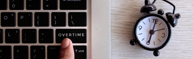 Imagem ilustrativa de um dedo pressionando a tecla “Overtime” em um teclado de notebook, ao lado de um despertador analógico, representando o controle de ponto, a jornada de trabalho e a gestão de horas extras nas empresas.